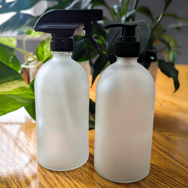 Two glass bottles with spray and pump tops on a wooden table in front of plants